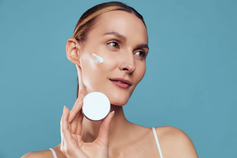 A young woman with blonde hair applies face moisturizer on her cheek, looking to the side, in a clean studio against a blue backdrop, conveying a fresh skincare routine and confident glow.