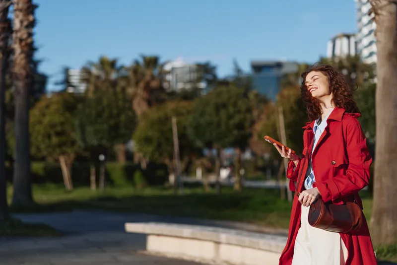 A stylish woman in a red coat stands in a sunny park, looking up with a relaxed smile, while palm trees and modern buildings frame the bright background.