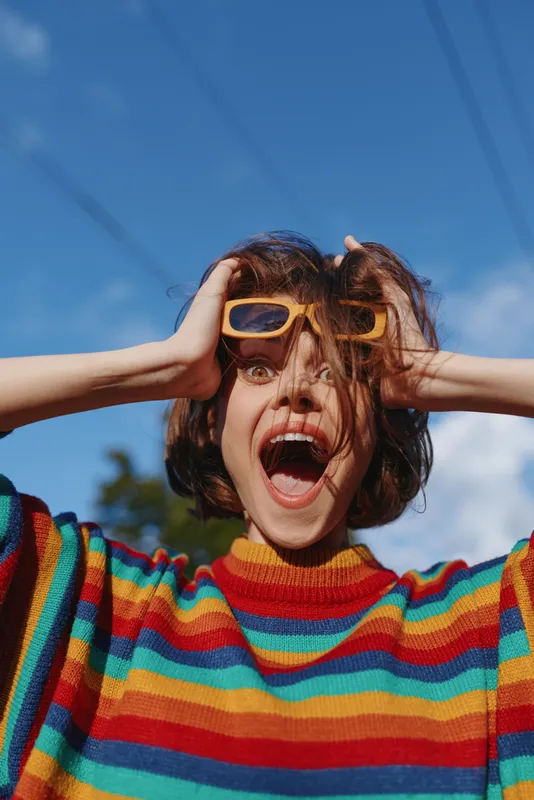 Young woman in a striped sweater wearing sunglasses with a surprised expression outdoors under a vivid blue sky, short brown hair, open mouth and joyful lifestyle fashion portrait.