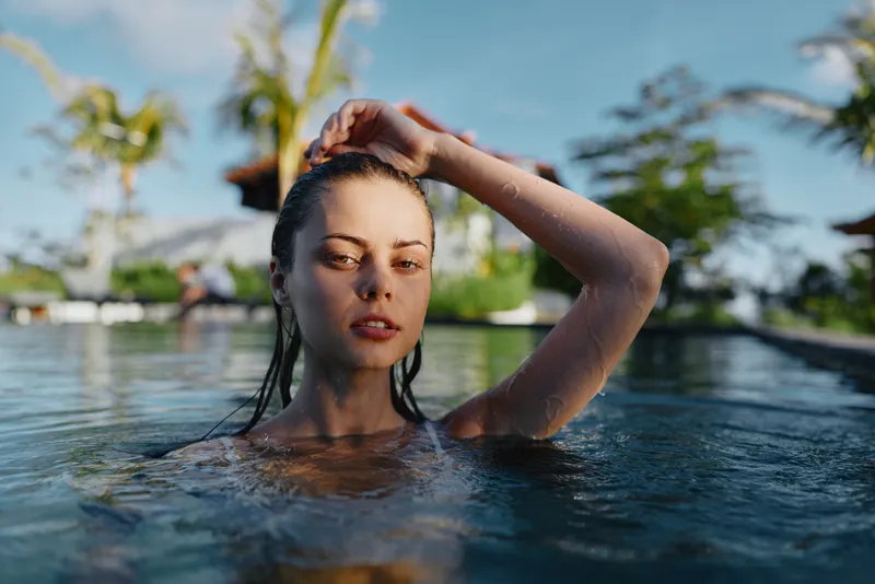 Young woman with wet hair relaxing in outdoor pool under natural sunlight. Concept of lifestyle, wellness, and summer leisure with tropical plants in background.