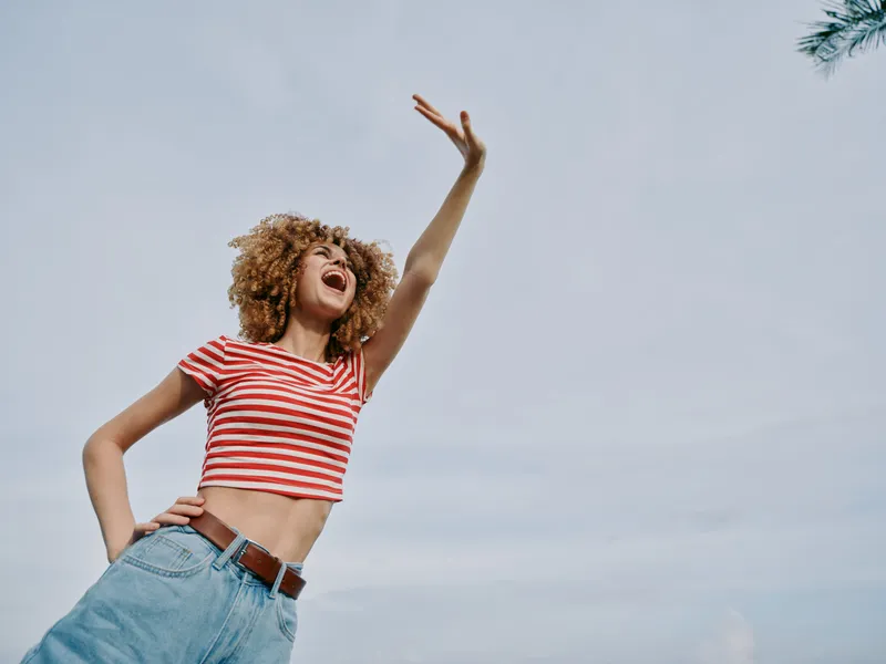 Energetic portrait of a joyful young woman outdoors, wearing a red striped crop top and denim jeans, posing mid motion with arm raised against a clear blue sky.