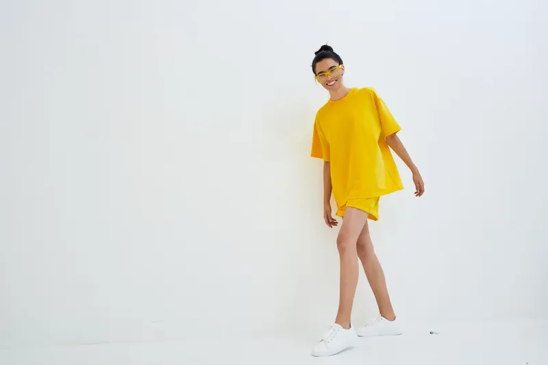 Young woman in yellow casual outfit walking confidently against white background, wearing white sneakers and yellow sunglasses, with a cheerful and relaxed vibe.