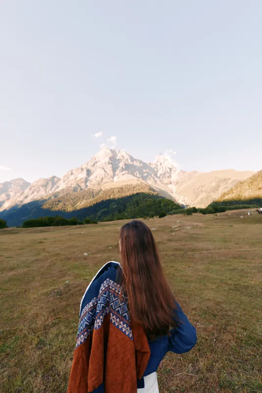 Woman with long hair from back wearing patterned shawl standing in meadow facing mountain landscape. Nature scene with wide grass field, outdoor travel and peaceful horizon.