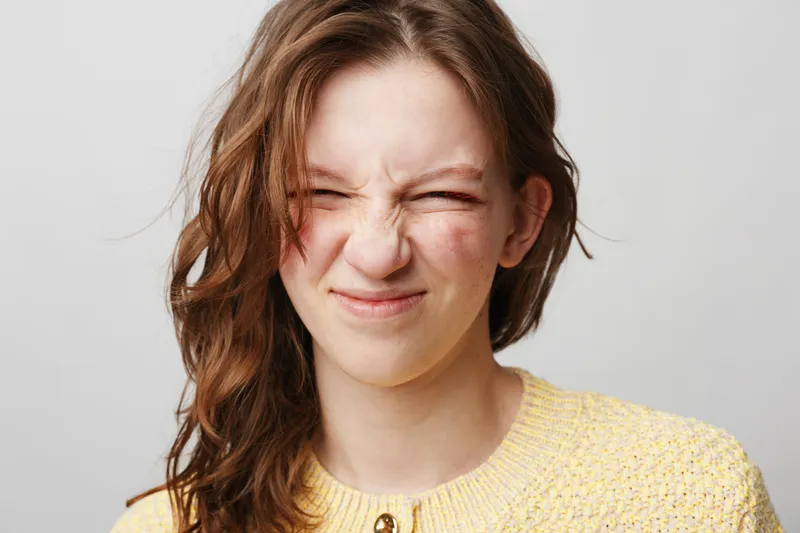 Young woman with long brown hair wearing yellow sweater makes a funny grimace with eyes closed. Isolated on white background. Concept of emotions, facial expression, and natural beauty.