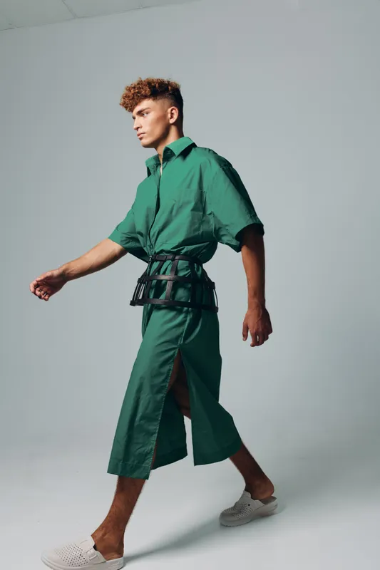 Man walking in studio wearing green dress with collar periodic slit and belt around waist modern fashion isolated on grey seamless background looking forward in profile.
