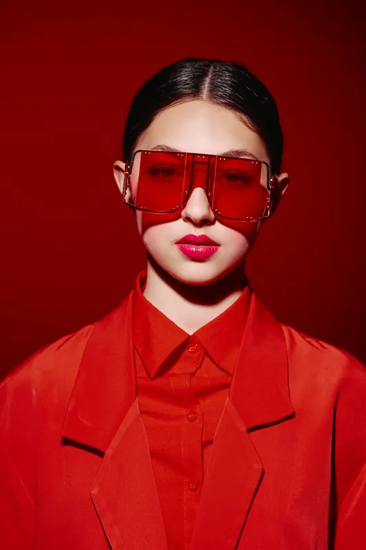 Fashion model posing with distinctive oversized red sunglasses and a stylish red blazer against a matching red background. Concept of beauty, creative style, and fashion.