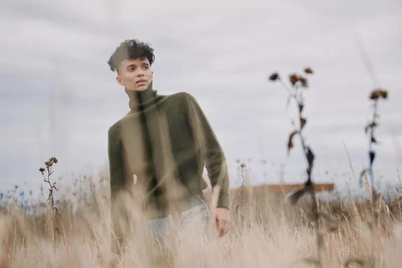 A young man stands in a windswept field wearing a turtleneck sweater, captured in a candid, contemplative portrait that conveys authenticity and credibility through natural light and pose.