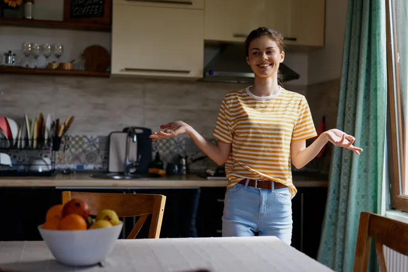 Young woman in striped t shirt stands in a bright kitchen, smiling with open hands, creating a casual home moment that feels warm and welcoming in a modern domestic setting.