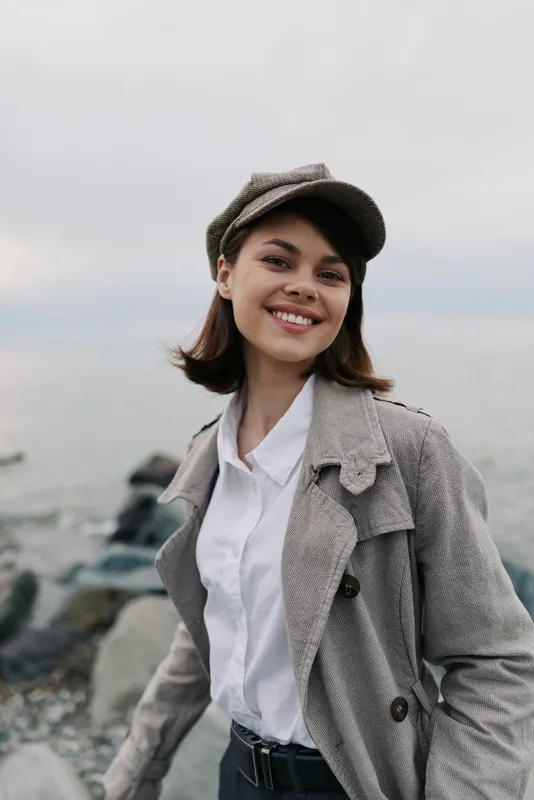 Smiling young woman wearing a textured coat and hat stands outdoors near a rocky shore. Soft natural light highlights her joyful expression and casual stylish outfit in cool weather.
