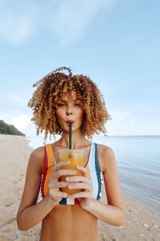 Woman with curly hair on beach, holding ocean juice with straw for summer hydration refreshment. Healthy lifestyle wellness concept. Bright sunlight, clear sky, calm waves.