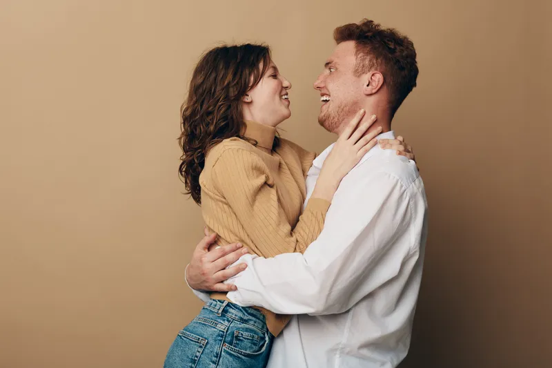 Happy young couple embraces and laughs together on beige background. Casual clothing, joyful mood, love and relationship concept with natural light and warm tones.