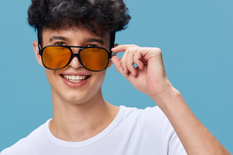 smiling man wearing yellow sunglasses and white t-shirt with curly hair on blue background, joyful young adult expressing happiness and style