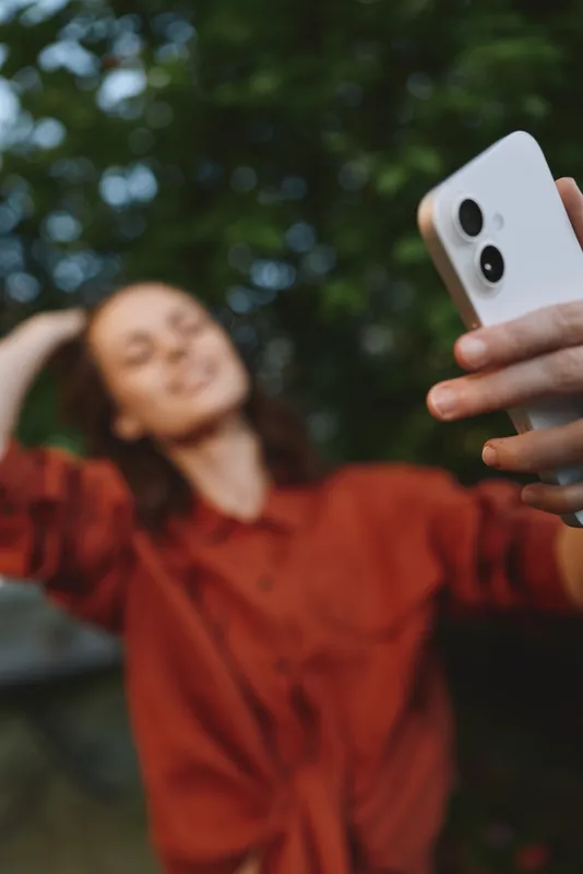 Happy woman lifestyle portrait outdoors, smiling while taking a selfie with smartphone in natural light, candid outdoor moment captured against greenery tones