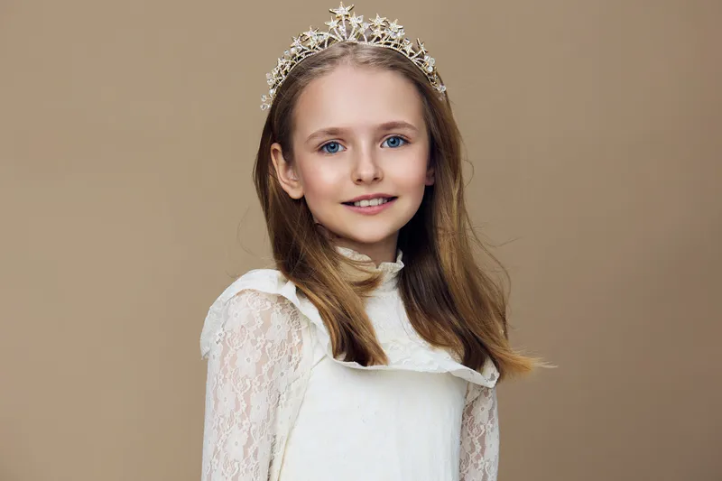 Young girl with a tiara smiling against a neutral background, showcasing elegance and charm in a light dress, perfect for celebrations and special occasions