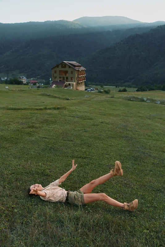 A woman lies on a green hillside, arms reaching up in a relaxed pose, wearing casual shorts and a light top. A rural house and distant mountains provide a peaceful landscape.