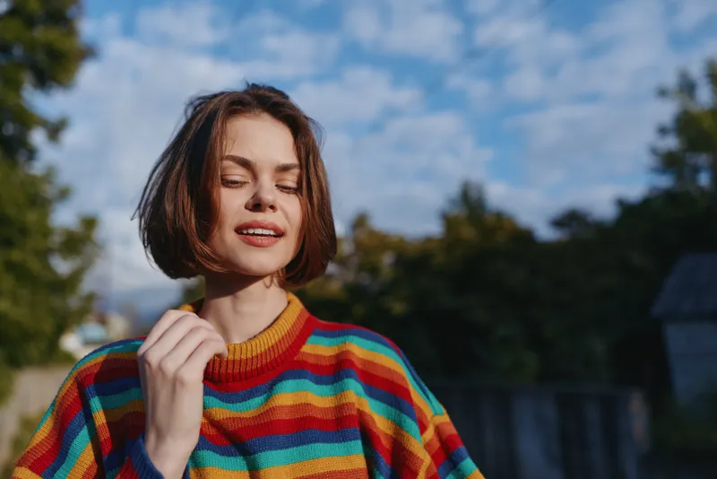 Woman portrait in colorful striped sweater outdoors, short hair and relaxed smile with closed eyes, natural sunlight on face, candid lifestyle shot with trees and blue sky.