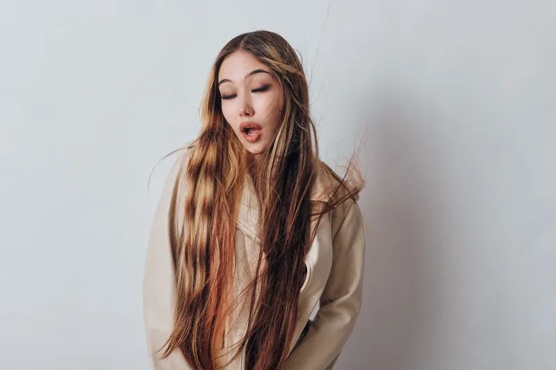 Woman with long hair and beige sweater making surprised facial expression on gray background. Portrait of female with flowing hair showing shock or amazement indoors in studio.