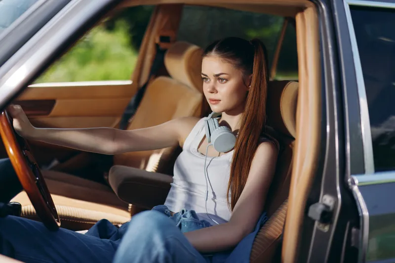 Young woman with long hair in casual attire, wearing headphones, sitting in a parked car during a sunny day, conveying a relaxed and carefree mood.