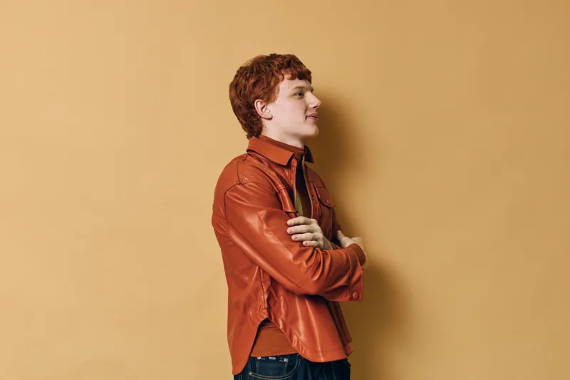 Young man with red hair wearing a brown leather jacket and jeans stands with arms crossed against beige background. Casual style, confident expression and side profile view.