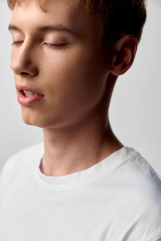 Young man with closed eyes, calm expression and soft lighting, wearing a white shirt, conveying serenity and focus in a minimal studio setting