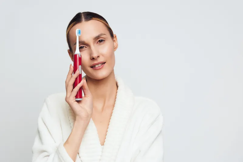 Young woman in a white bathrobe holds a toothbrush near her face, smiling softly in a bright studio, conveying skincare, self care and wellness with a calm, intimate mood, with a clear focus and