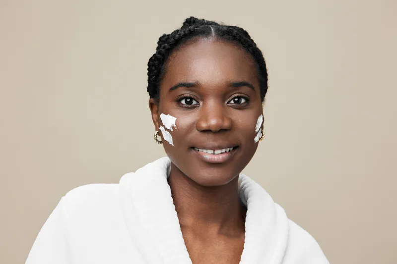 Portrait of a smiling black woman with skincare cream on her face wearing a robe, promoting self care and beauty in a neutral toned background