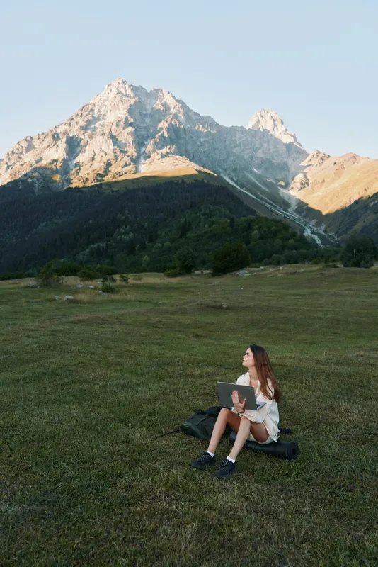 A young student sits on a grassy field in a mountainous landscape, using a laptop, focused and calm as sunlight bathes the scene with natural serenity.