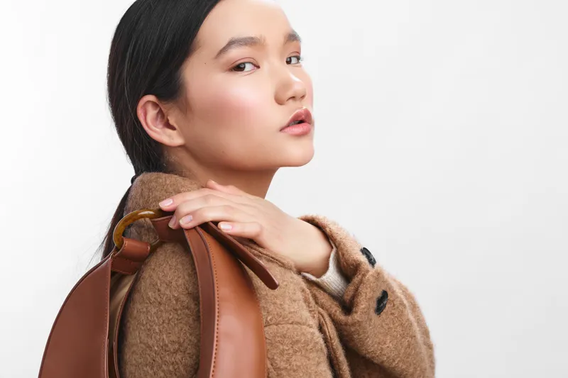 Asian woman stands in a clean studio wearing a brown coat and backpack, projecting calm confidence and contemporary style. The simple backdrop emphasizes fashion and everyday life.