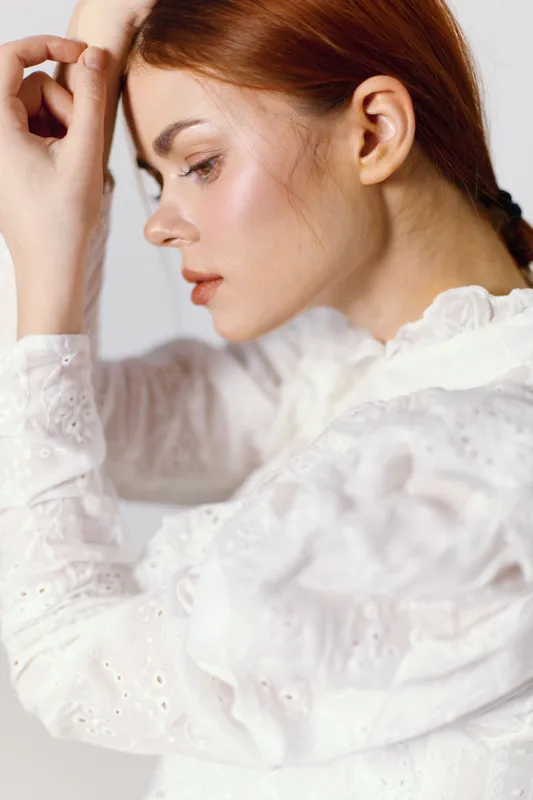Profile portrait of a young woman with red hair in a white blouse, conveying a contemplative emotion against a light background, perfect for wellness or beauty themes