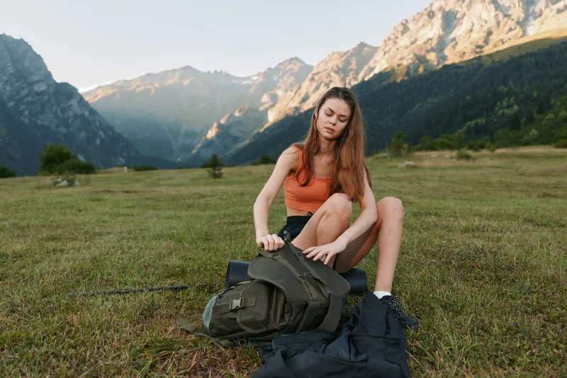 A young woman outdoors in a grassy meadow prepares gear with distant mountains. She sits, unpacks a backpack, and concentrates with a calm, focused expression.