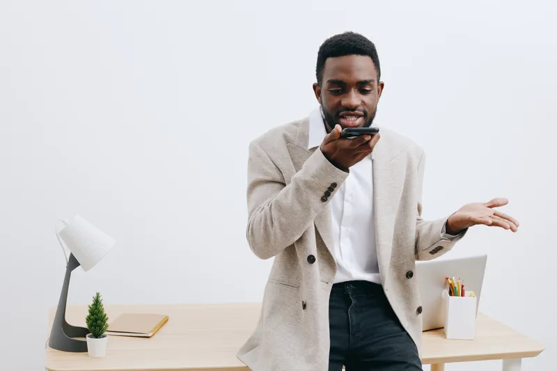 Casual man using smartphone in modern office interior, standing near desk with stationery, potted plant, and lamp, professional workspace, skilled worker, communication concept.