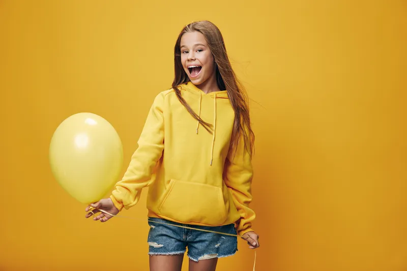 Girl in yellow hoodie holding yellow balloon smiling and looking away on bright yellow background. Casual young female in denim shorts posing with party decoration for celebration.