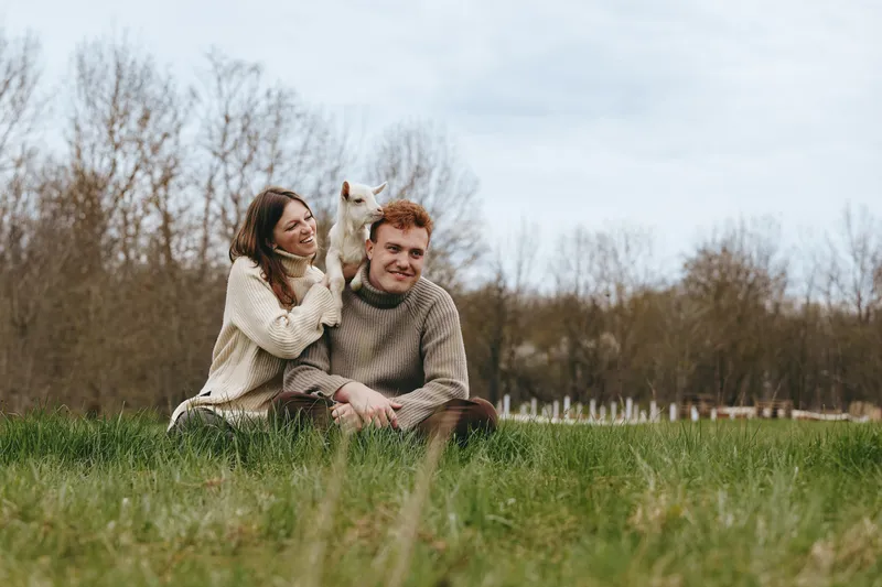 Casual couple outdoors with a playful goat kid, sitting on grass. Warm, candid lifestyle scene highlights companionship, nature, and farm charm in soft daylight.