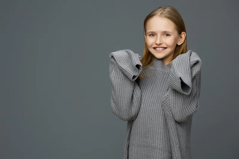 Young girl smiling in a cozy gray sweater with oversized sleeves against a neutral background, conveying warmth and happiness during autumn