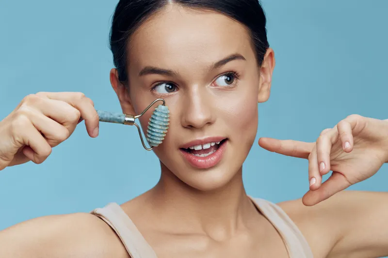 Young woman using a jade roller on her face, showcasing a fresh and glowing complexion against a pastel blue background Emphasizing self care and beauty routines