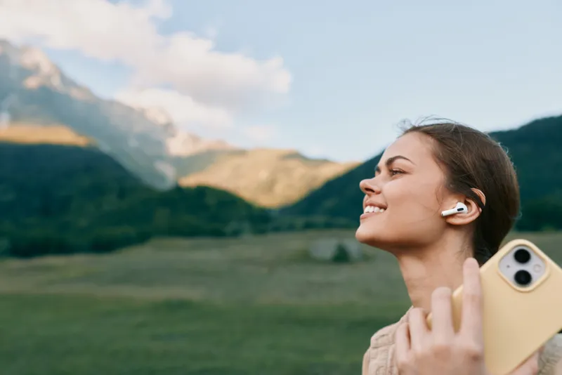 Young woman smiles in a sunlit mountain landscape as she takes a casual selfie with wireless headphones, enjoying fresh air and open space.