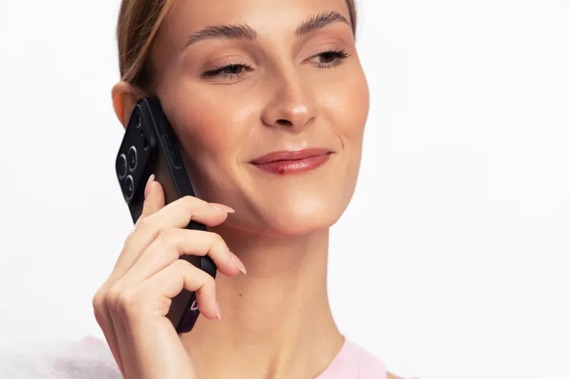 Young woman with clear skin holds black smartphone to ear, smiling slightly against a white background. Concept for communication, technology, and modern lifestyle with positive mood.