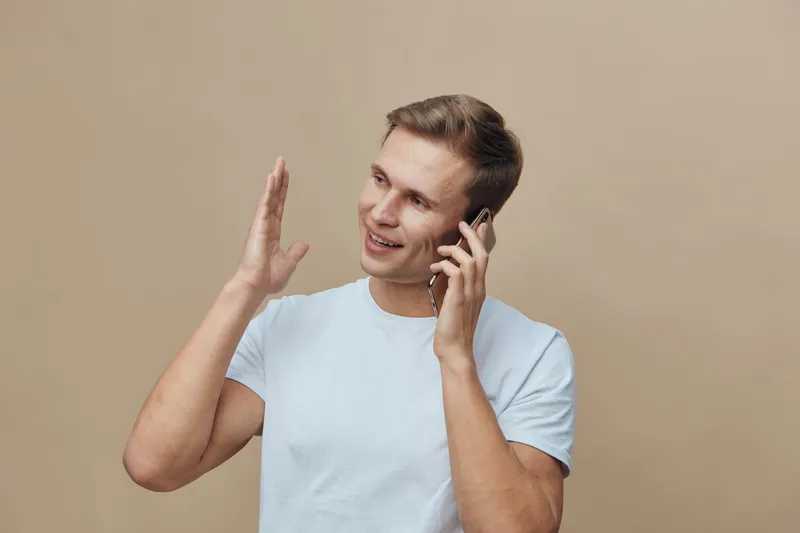 Smiling man talking on mobile phone with cheerful expression, casual light blue t-shirt, beige background, indoor setting, lifestyle communication, happiness, technology.