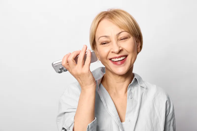 Smiling senior woman holding a smartphone near her ear, having a phone call, focused portrait on a clean light background, casual indoor communication, warm expression, cheerful