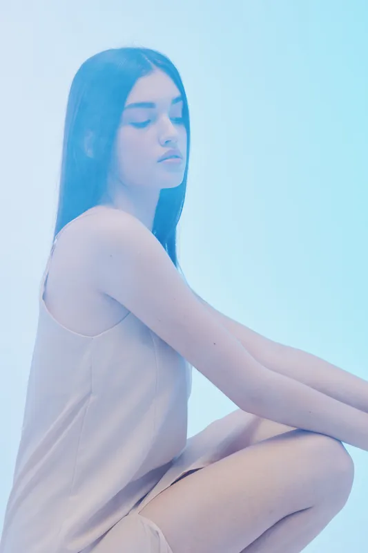 Young woman with long dark hair poses in a soft pink dress against a light blue background. Concept of beauty, fashion, elegance, and calmness in studio lighting.