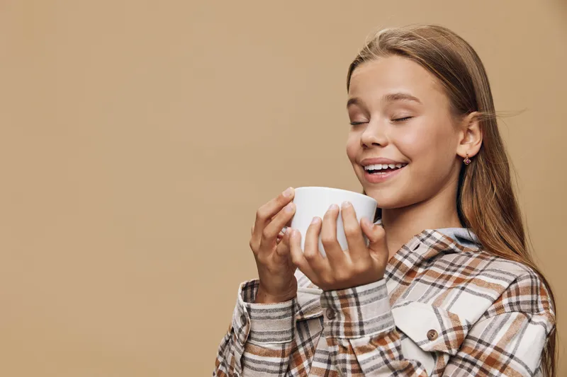 Girl holding white cup with closed eyes enjoying hot drink in cozy plaid shirt against beige background. Relaxation, comfort, warm beverage, casual style, peaceful moment with happiness.