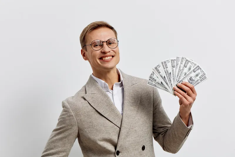 Smiling confident businesswoman with short hair and glasses holding fanned-out stacks of hundred-dollar bills, dressed in smart beige blazer, white shirt, posing against plain white background, studio