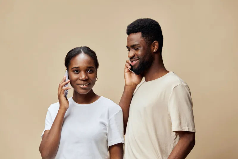 Two young African adults engaged in a joyful conversation on mobile phones against a neutral beige background, expressing happiness and connection