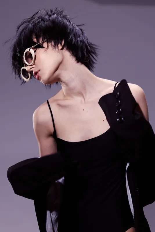 Stylish woman with short hair wearing unique round sunglasses and black outfit, posing against a soft purple background in artistic fashion style.