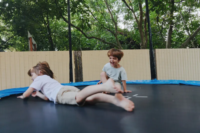 Two happy children playing on a trampoline, enjoying their carefree moments outdoors in a lively green setting with sunlight filtering through the trees.