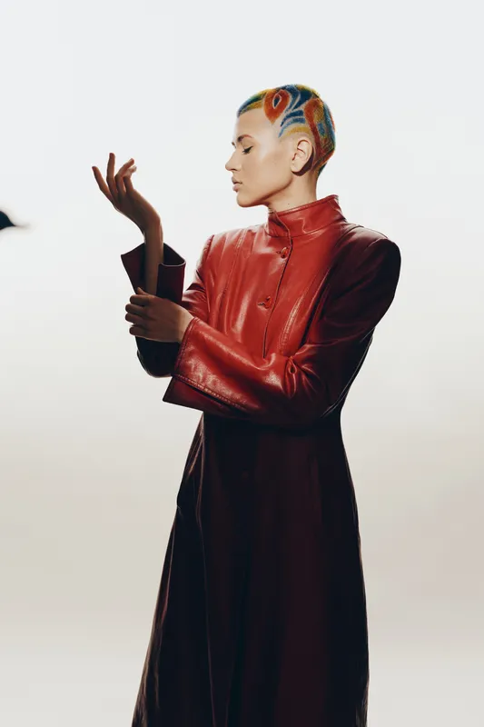 Stylish woman with colorful braided hair wearing a red leather coat, posing thoughtfully in a minimalistic studio with soft lighting and neutral background.