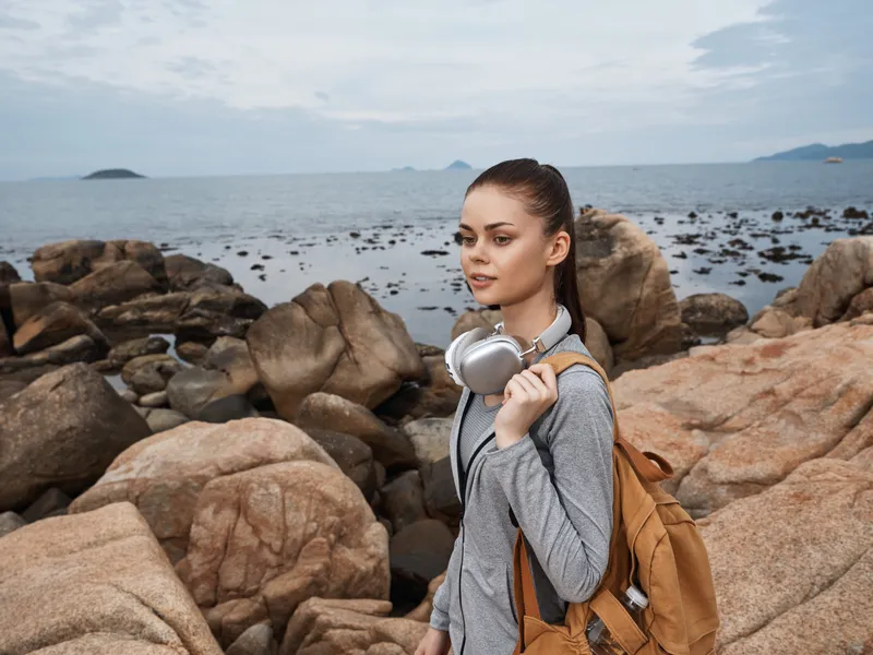 Young woman in casual gray outfit with headphones, standing on rocky shore by the sea, showcasing a relaxed and adventurous spirit