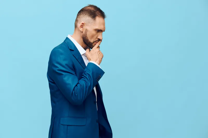 A professional man in a blue suit stands against a solid blue backdrop, conveying deep thought and focus as he contemplates a decision.