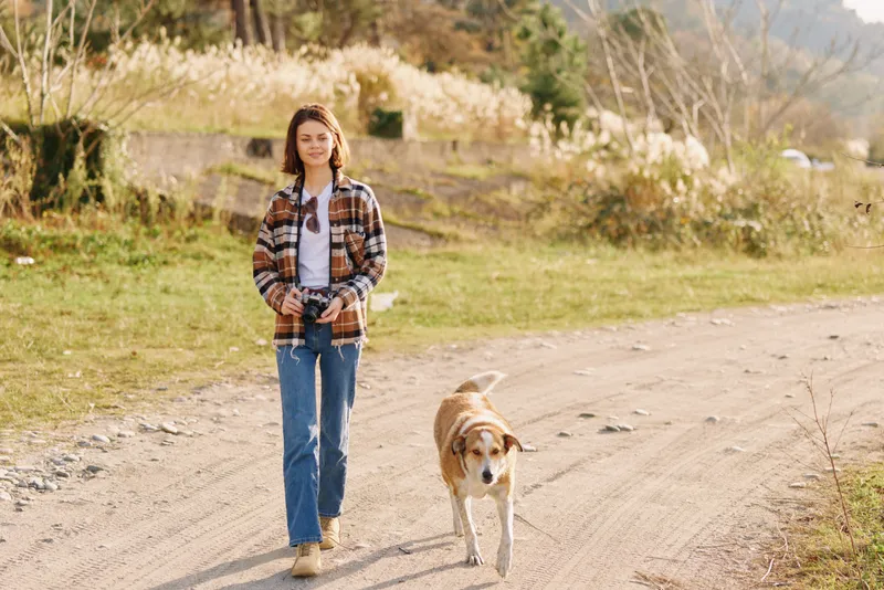 Woman walking with dog along a dirt path in a sunny outdoor setting, wearing a plaid shirt and jeans, capturing a peaceful nature moment on the trail.
