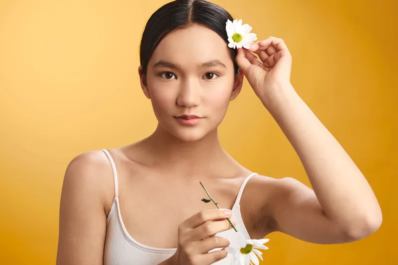 Asian woman holds daisies against a warm backdrop, with soft lighting highlighting her calm expression and natural beauty, creating a fresh, serene portrait suitable for lifestyle or wellness